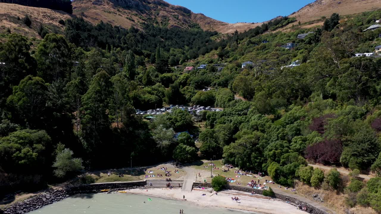 Drone shot moving away from bay with people on beach, swimming and pine trees and houses on hill in background