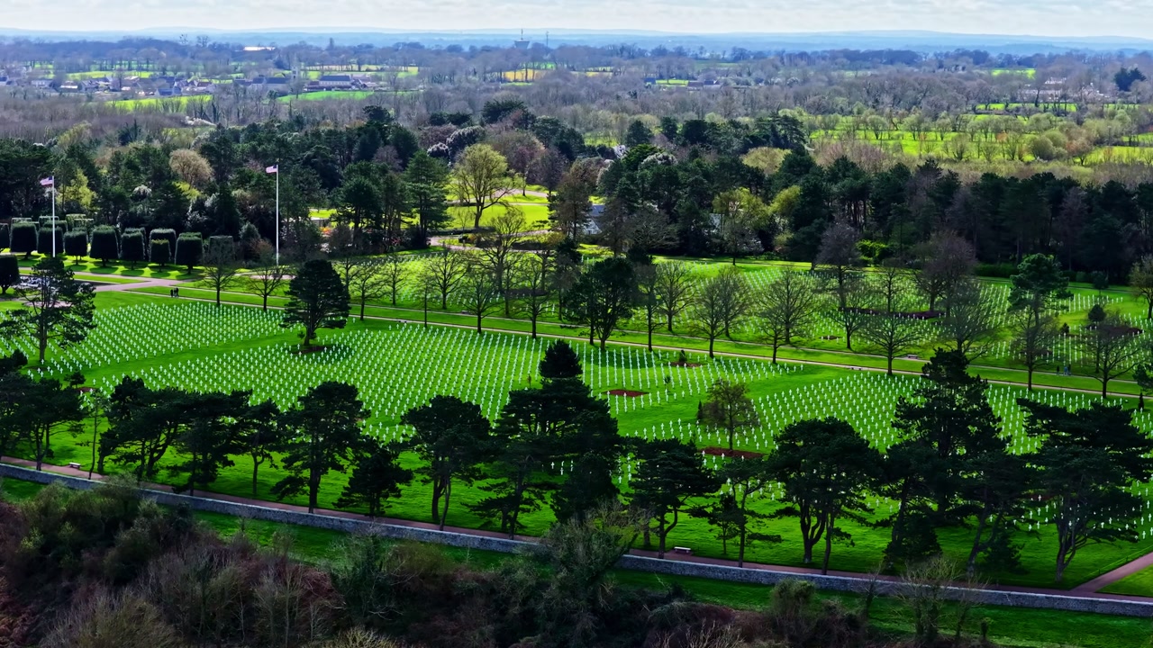 Aerial drone flyover reveals thousands of white crosses in rows at an American WWII military cemetery surrounded by lush green trees in Herbignac, France