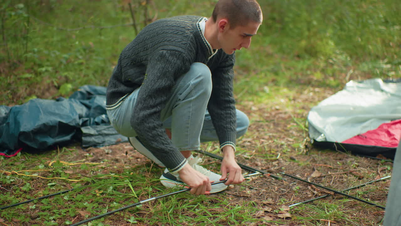 Man in gray sweater crouches on forest ground assembling tent poles beside open tent fabric while woman offers assistance, surrounded by camping supplies and green woodland background
