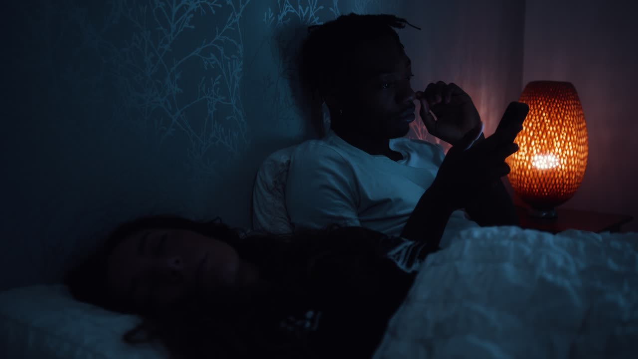 男性は夜にベッドに座ってスマートフォンを使い疲れから目を擦ります