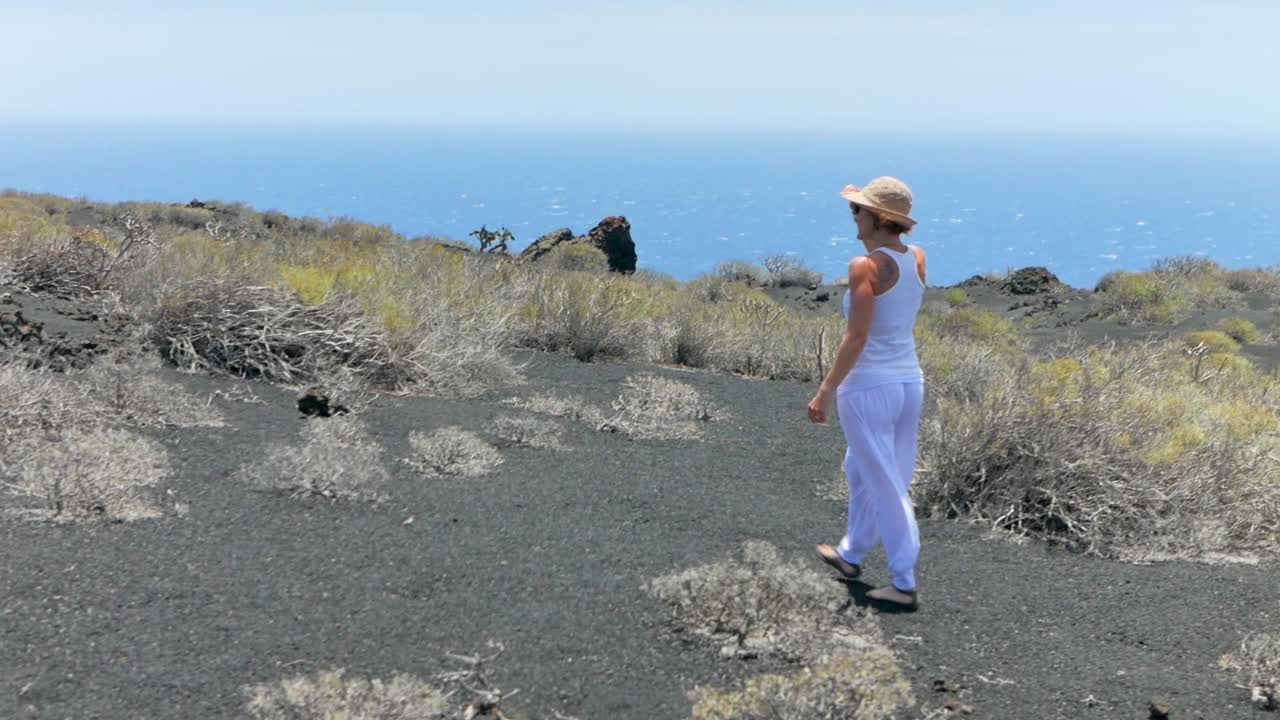 A shot following an elegant woman in white walking on a volcanic island, and losing her hat a cause of wind