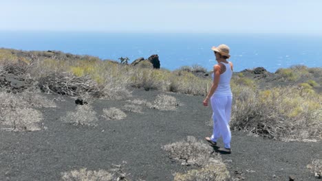 a shot following an elegant woman in white walking on a volcanic island, and losing her hat a cause of wind