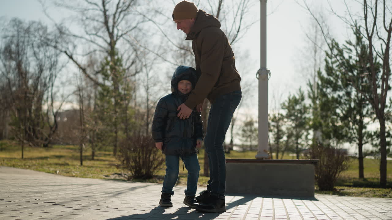 un primer plano de un padre levantando a su hijo ambos están vestidos con vaqueros y botas, con el niño con una chaqueta hinchada y el hombre con un gorro, árboles y un poste eléctrico están alrededor