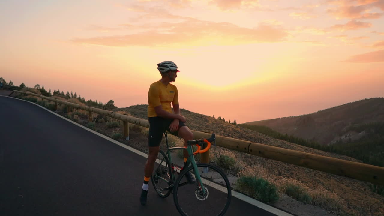 Sports enthusiast in yellow t-shirt, helmet, and gear relaxes on mountain bike, taking in mountains and setting sun after training