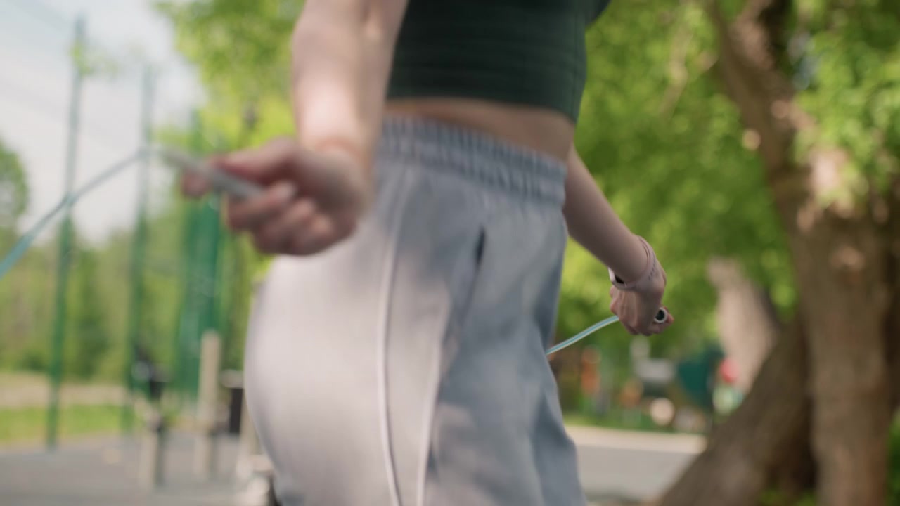 Close up of young woman actively skipping rope in outdoor park setting wearing gray sweatpants and holding jump rope handles. background features green trees distant playground equipment