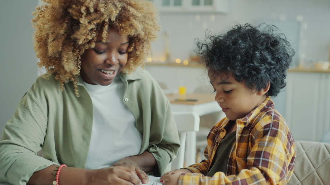 African American Mother Talking to Cute Little Son Drawing on Paper at Table