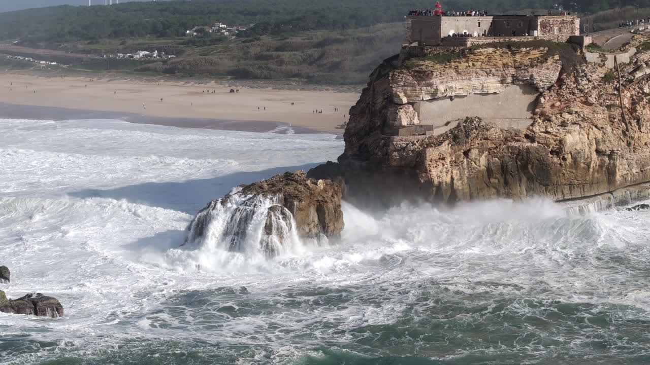 Professional aerial drone shot of big waves smashing into and breaking on rocks and cliffs in Nazaré, Portugal, Europe. Farol da Nazare lighthouse and wind turbines visible