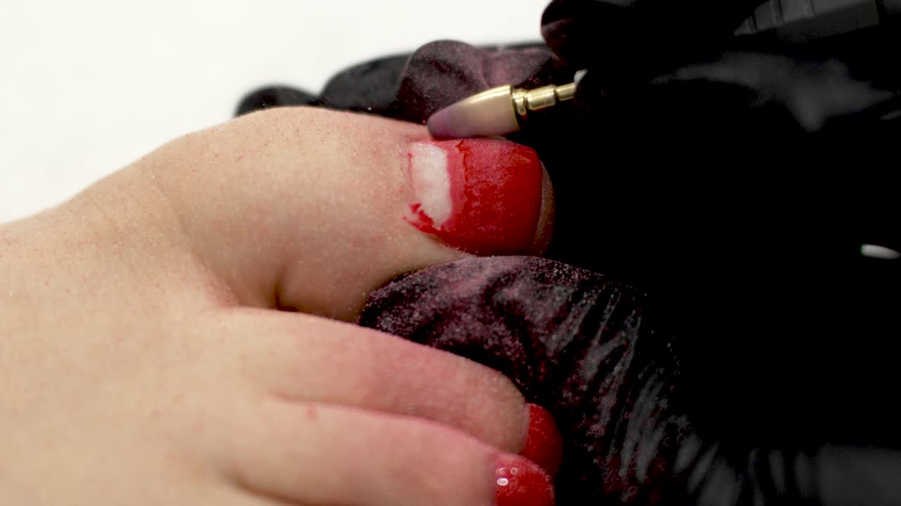 Manicurist nail technician in black gloves removes red gel polish on big toe using a nail drill, performs a pedicure for woman client