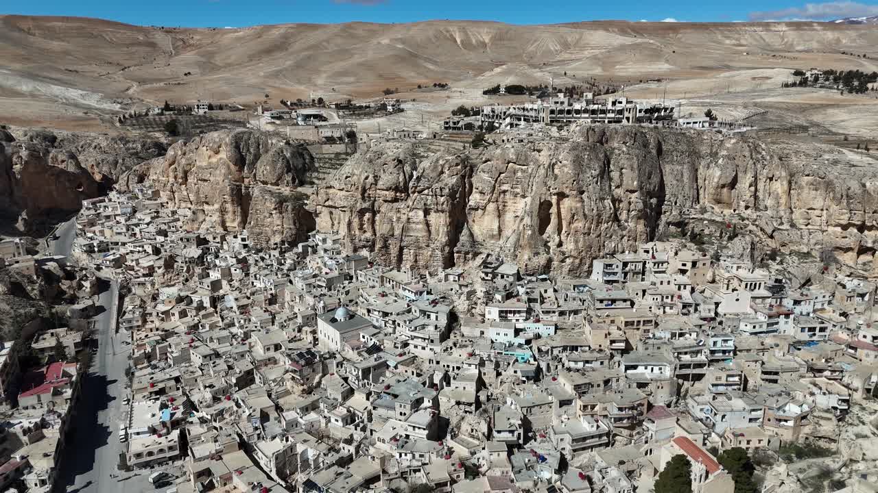 A wide aerial drone panorama of Ma'loula, Syria, showcasing the ancient village built into the massive rocky cliffs of the Qalamoun Mountains