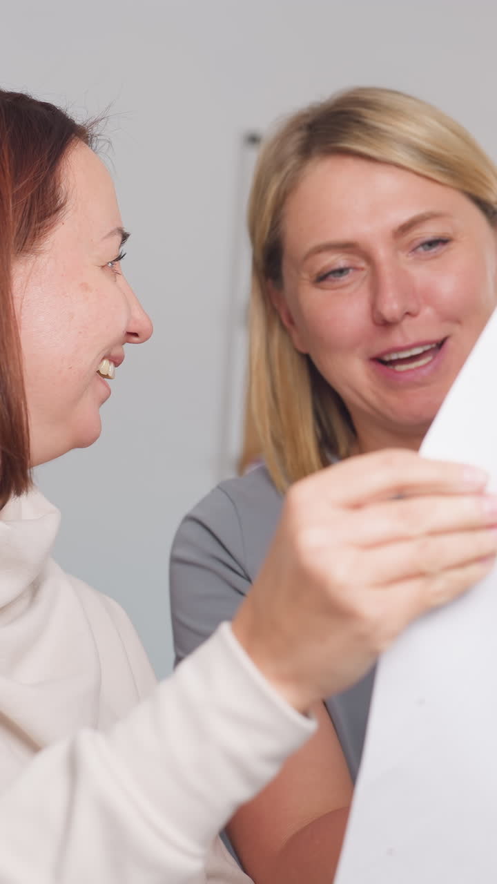 Dos mujeres blancas inspeccionan una muestra y el ajuste en un estudio luminoso, la responsable de producto comprueba las marcas mientras la patronista explica los ajustes; revisión práctica de calidad y aprobación en colaboración