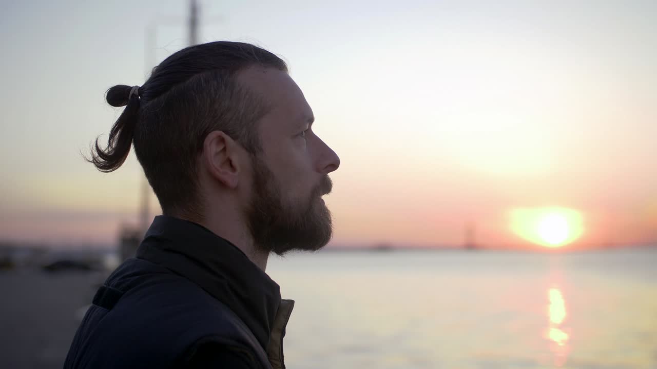 retrato de un elegante hombre hipster en el fondo del puerto marítimo durante la puesta de sol, mirando a un lado