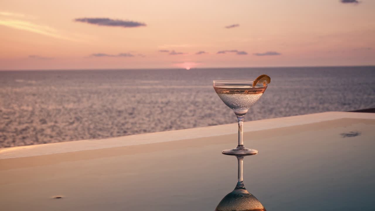 Elegant cocktail glass with condensation and orange peel is standing on the edge of an infinity pool's wet surface, reflecting the beautiful sunset over the calm ocean
