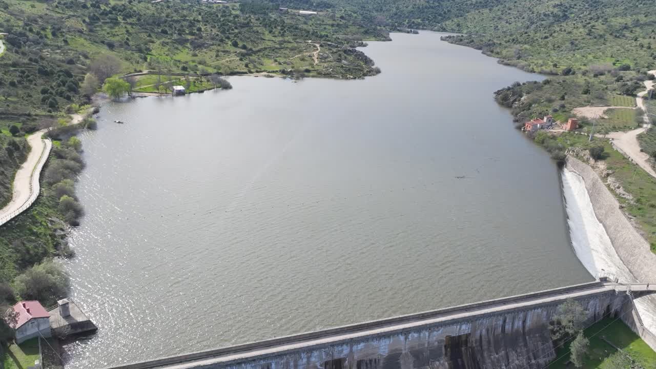 Ascending drone flight over a dam with a descending camera pan showing the retaining wall, side spillway releasing water, and the impressive size of the reservoir and its scenic surroundings.