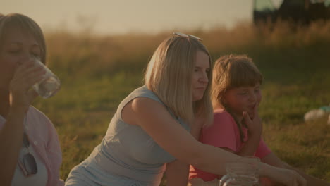 partial view of middle-aged woman drinking water outdoors while seated next to mother and daughter, mother is reaching for green grapes and eating it, and daughter is smiling