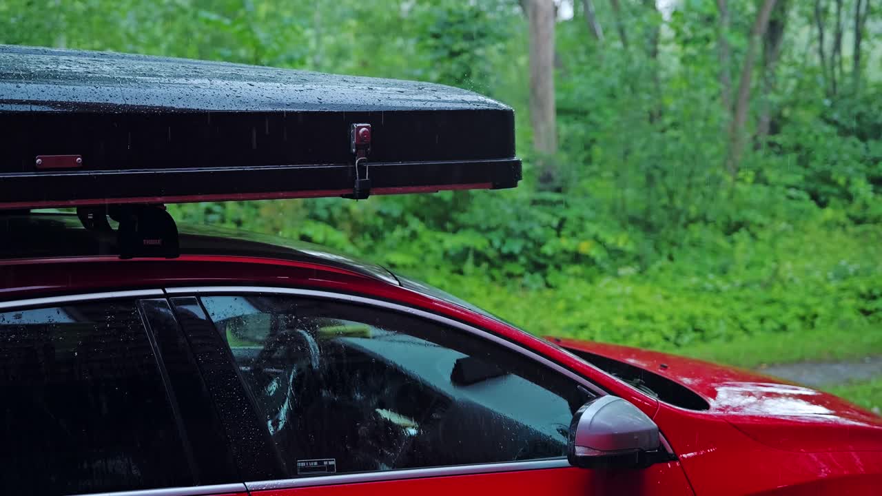 Rain pours heavily on red car with rooftop box in forest slow motion shot