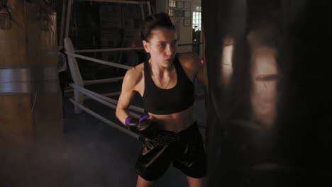 Slow-Motion-shot-of-a-female-boxer-in-gloves-hitting-hard-a-boxing-bag-with-her-fist-while-training-in-a-dark-fitness-studio-with