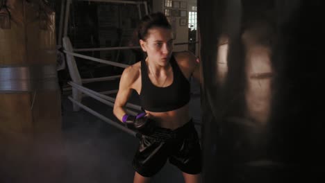 slow motion shot of a female boxer in gloves hitting hard a boxing bag with her fist while training in a dark fitness studio with