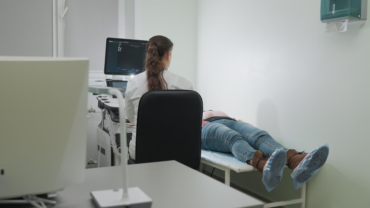 Medical doctor prepares for patient examination by retrieving scanner tool and placing it on faceup patient in clinical setting with diagnostic machine, desktop computer, and lamp stand visible