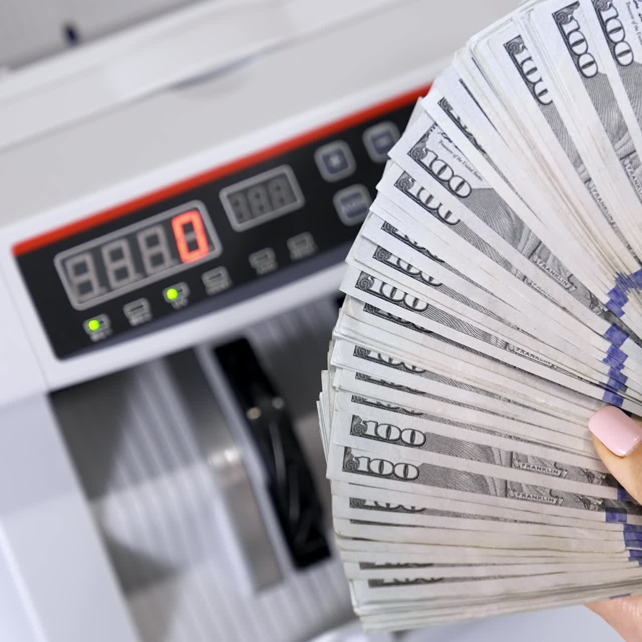 Bundt af hundrede dollarsedler. Kvindes hænder holder en stor sum dollarsedler med en automatisk tællemaskine i baggrunden.