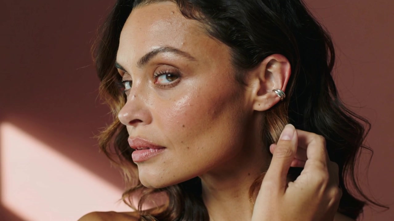Modella che alza la mano destra, sistema un ear cuff, si sistema i capelli per un primo piano in studio, spalle scoperte, anelli