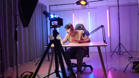 asian man holding mobile phone and read comment in laptop computer then talking to camera while live stream, professional light equipment