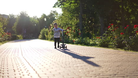 cute smiling baby boy turns his scooter on the paved road in the park. kid walks approaching camera and then rides pushing from the ground.
