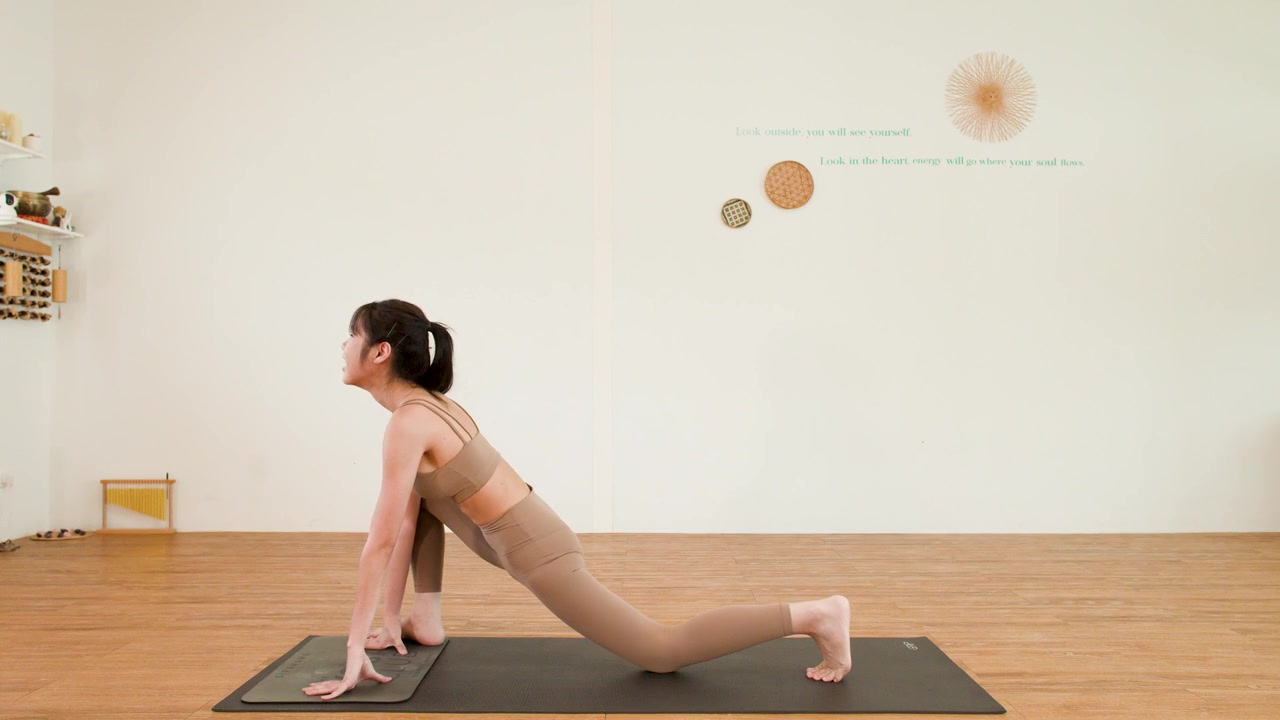 A woman in beige activewear practices a low lunge on a black yoga mat