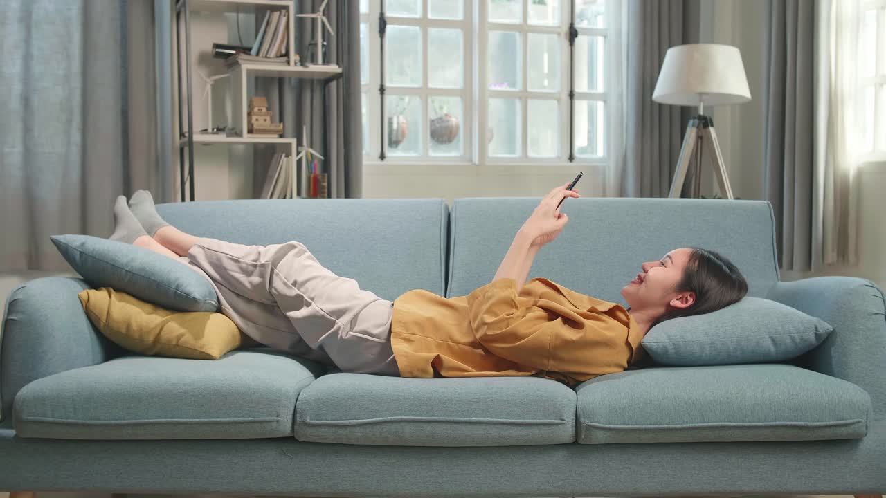 Asian Woman Lying On Couch In Living Room While Using Smartphone