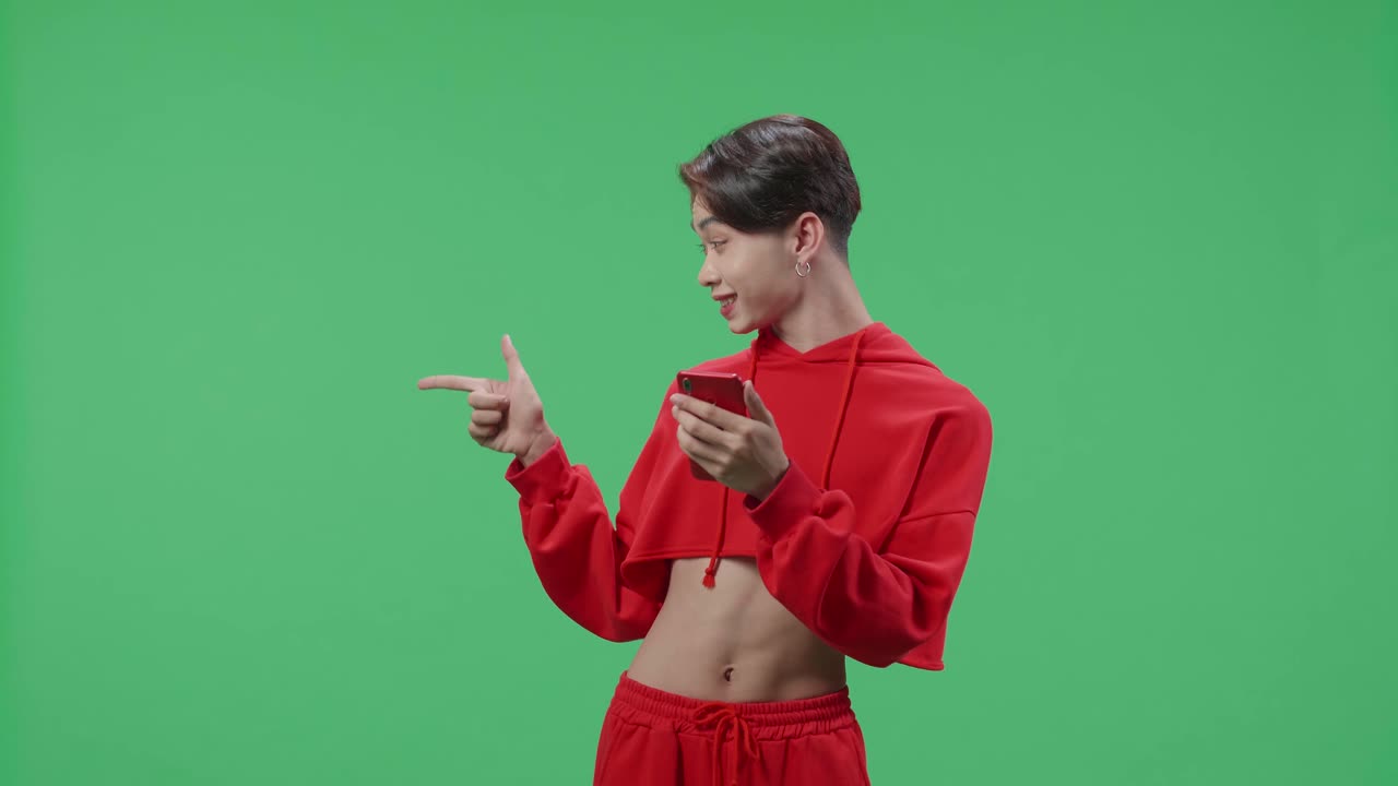 The Inspired Asian Transgender Male Flipping Through The Mobile Phone With A Smile Pointing To The Side While Standing In The Green Screen Studio