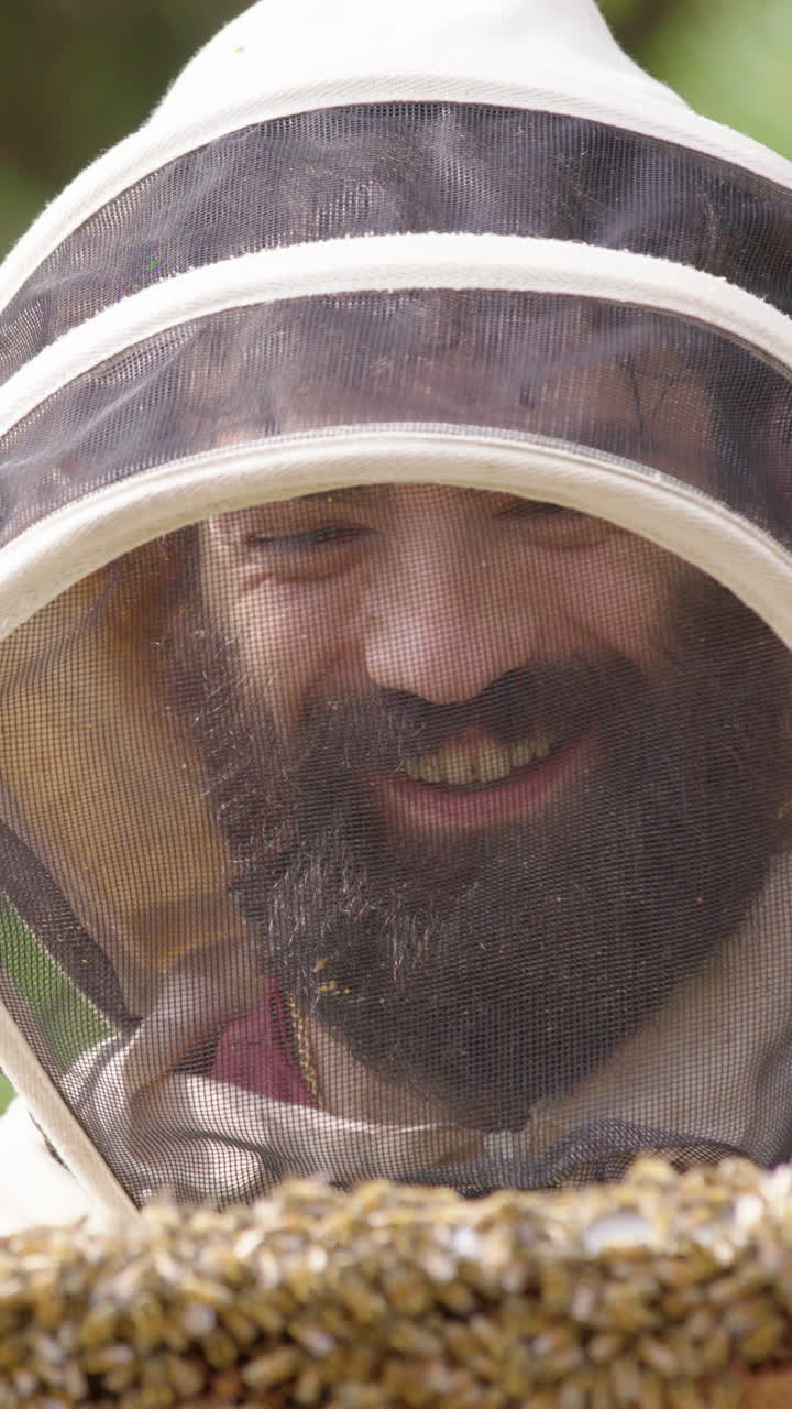 Happy beekeeper in bee suit inspects hive frame covered in honeybees, vertical