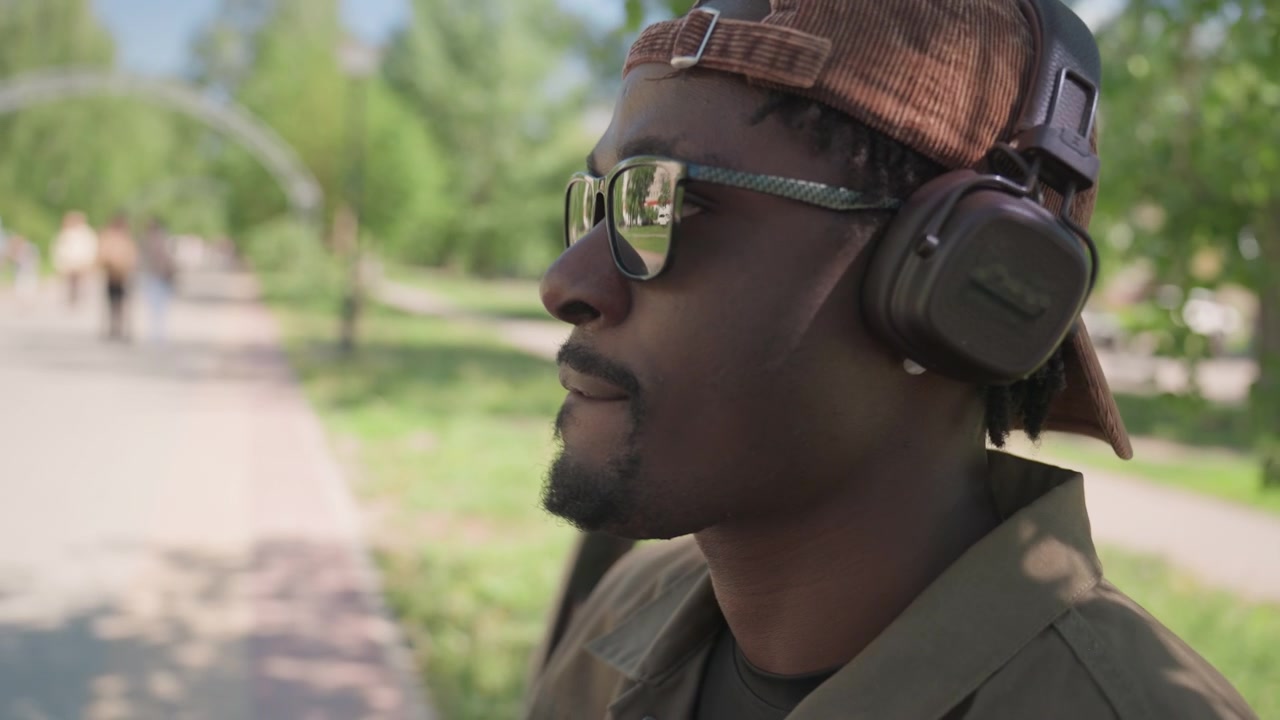 Close up of man wearing brown cap and headphones looking out car window at park scene with green trees and people walking. Man remains in profile view throughout showing consistent