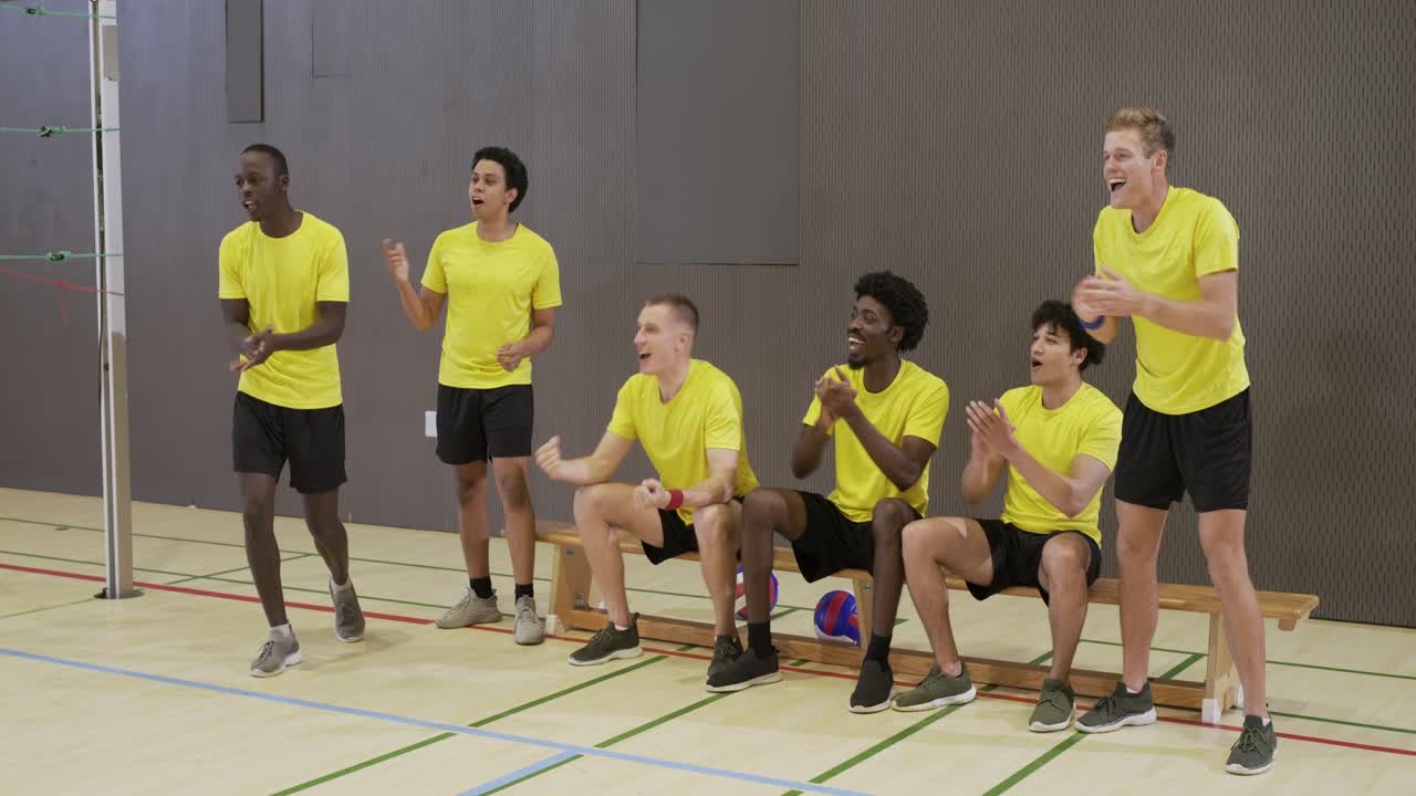 Sechs unterschiedliche männliche Volleyball-Teamkollegen in gelben Trikots feiern einen Punkt auf einer Turnbank mit Ball.