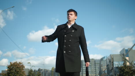 banker in black coat retrieving phone from pocket to answer call while walking confidently outdoors in urban setting with modern buildings and greenery visible in blurred background