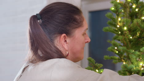 Side view of woman in brown wear carefully arranging Christmas lights on festive tree indoors creating warm cozy holiday atmosphere with gentle lights and joyful seasonal preparation mood