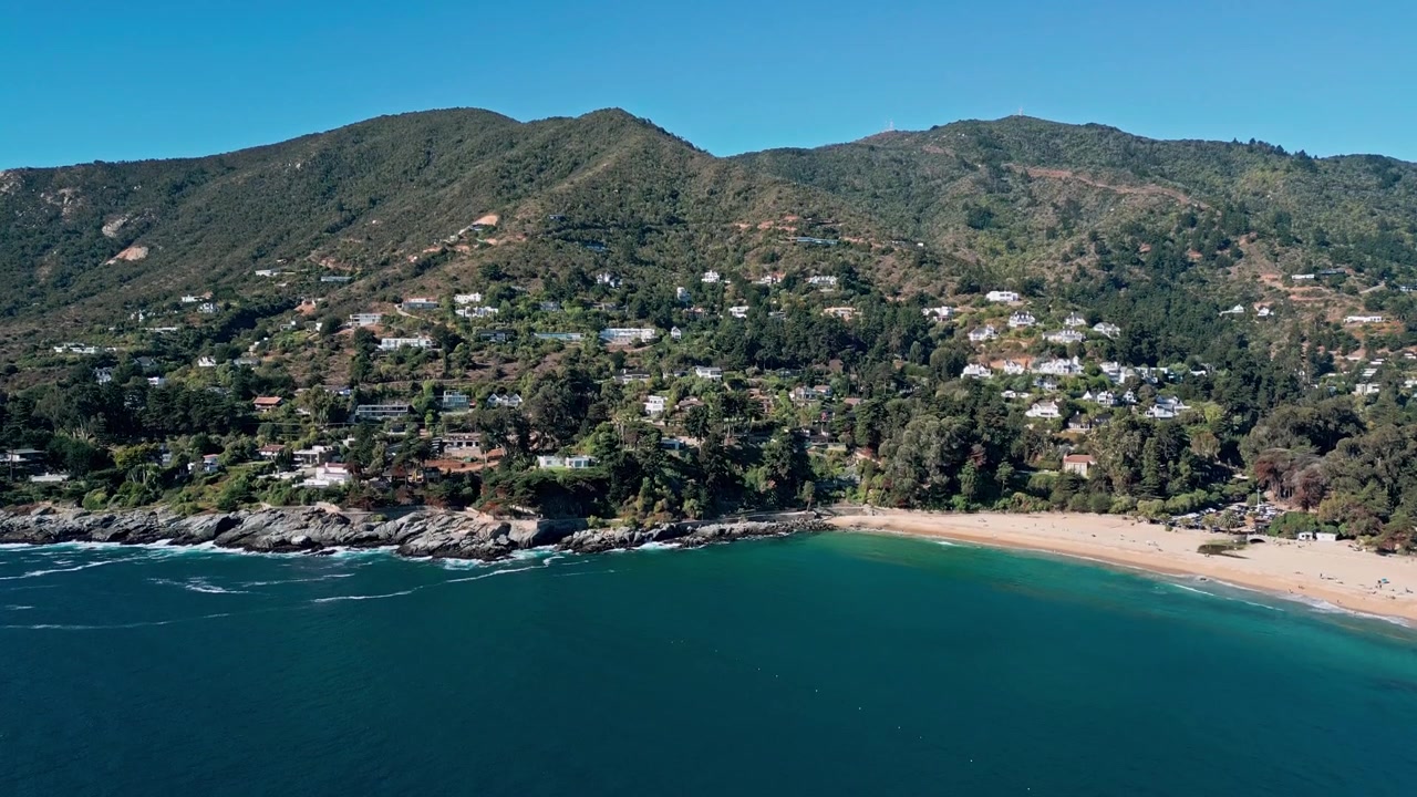 Toma descendente sobre una bahía azul profundo hacia una costa rocosa con villas exclusivas escondidas entre el bosque nativo que descienden hasta la cala
