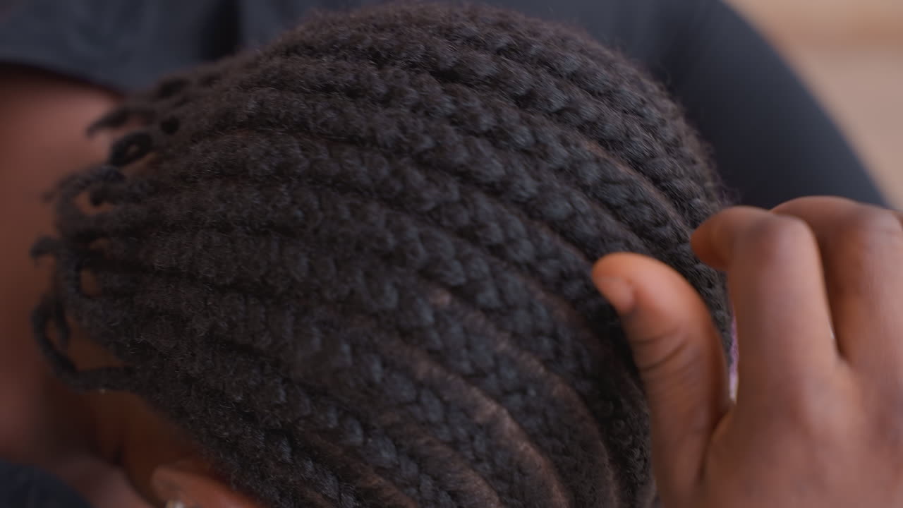 Vue de dessus d'une femme noire terminant des tresses africaines traditionnelles, mains lissant les extrémités en rangées symétriques. Ambiance de cérémonie culturelle, lumière du jour chaude, cadre communautaire intime, style ancestral, soin et célébration.