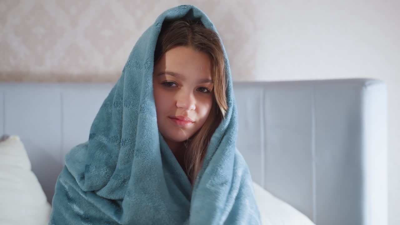 Young girl sits wrapped in soft blue blanket with hood over head, smiling playfully while sitting on bed in cozy room with neutral wallpaper and pillows, suggesting warmth, comfort, and relaxation