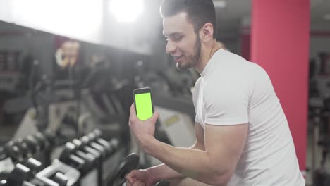 While-other-people-work-out-at-back,-a-sportsman-stands-near-mirror,-holds-a-dumbbell-in-one-hand-and-his-phone-in-other-hand-facing-camera