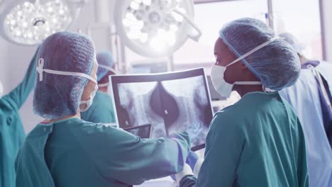 Video-Der-Rückansicht-Zweier-Unterschiedlicher-Chirurginnen,-Die-Im-Operationssaal-über-Röntgenaufnahmen-Diskutieren