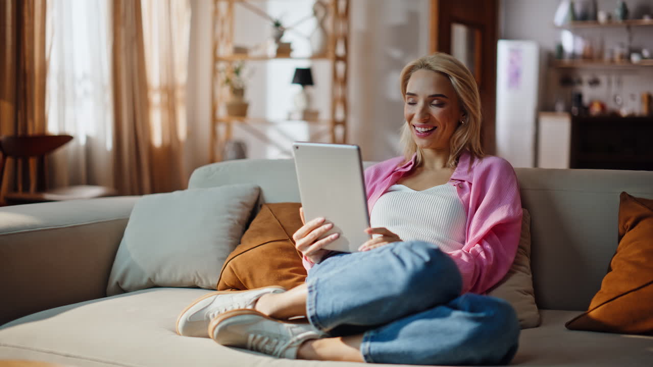 Home woman touching tablet screen at grey sofa. Laughing blonde using pad online