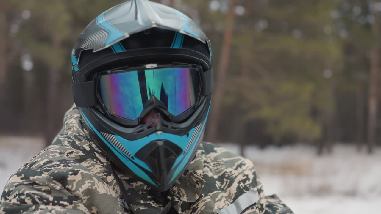 Helmeted rider in snowy pine background stares through mirrored goggles, teal helmet and camouflage jacket, breath visible in cold air, focused expression, quiet preride pause, reflection of trees