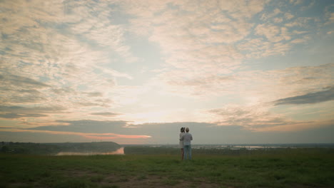 a husband and wife stand on a grassy hill beside a lake at sunset, gazing at the sky. the man, in a white shirt, hat, and jeans, holds the woman by the waist. she wears a black hat and a white dress