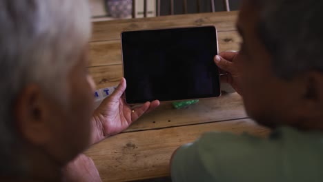 Senior-mixed-race-couple-using-tablet-talking-at-kitchen-table