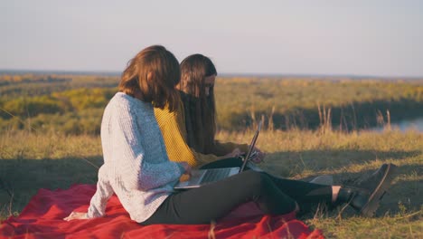 women use tablet and laptop sitting on red plaid in camp