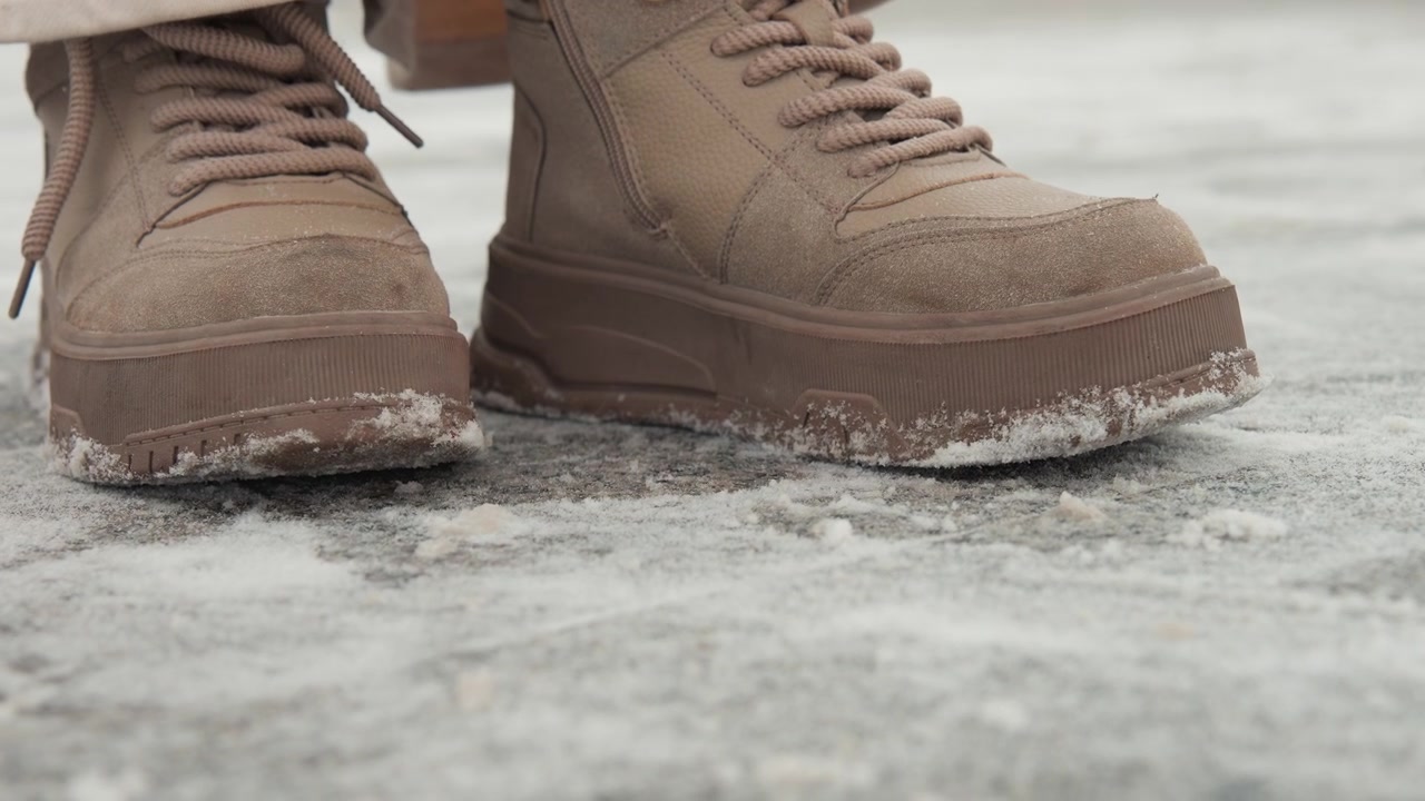 Low-angle close-up brown lace-up winter boots on textured icy ground with frost on sole edges and fabric, while loose laces drape over frozen surface and careful movement suggests winter footing