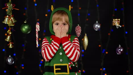 Kid-girl-in-Christmas-elf-Santa-Claus-helper-costume-looks-at-camera-makes-big-eyes-covering-mouth