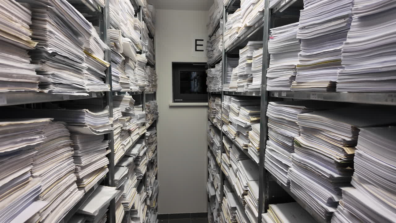 A narrow archive room filled with tall metal shelving units stacked with thousands of paper documents and files, creating dense rows of records on both sides