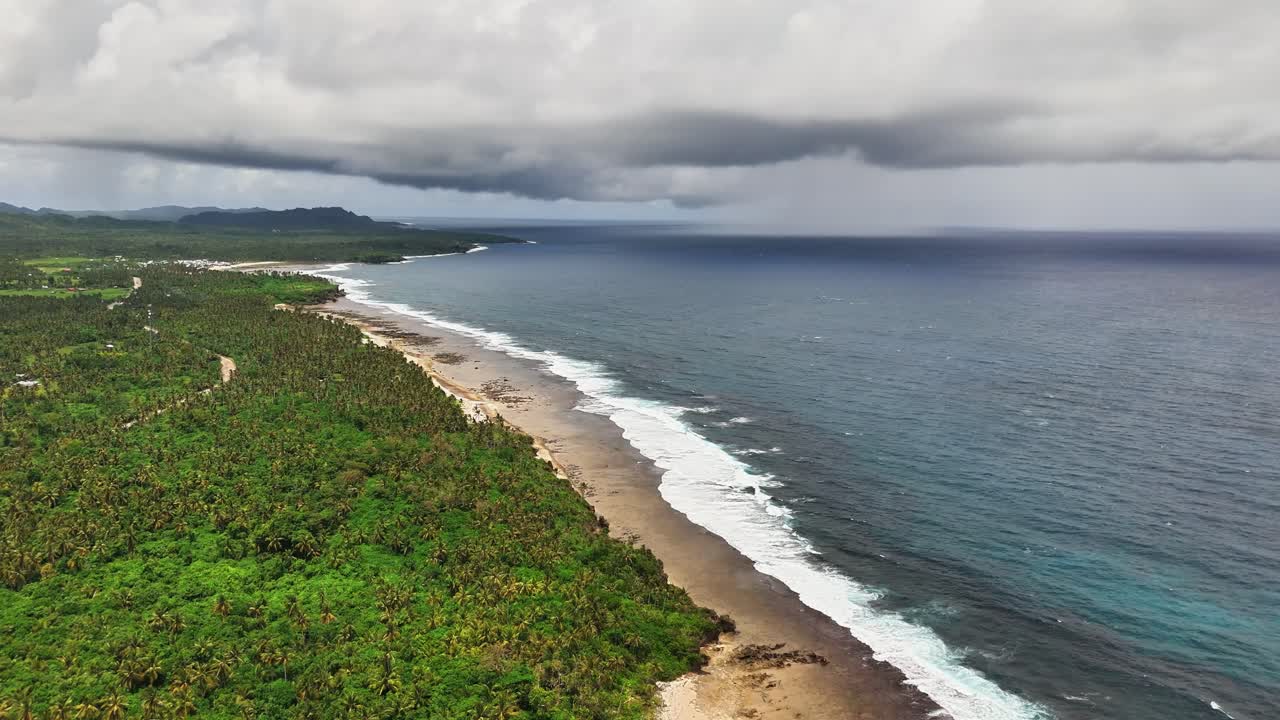 Drone shot over Magpupungko, Siargao Islands, Philippines showing coconut forest, sandy beach and waves of the Philippine Sea under a cloudy sky in a remote tropical coastal environment