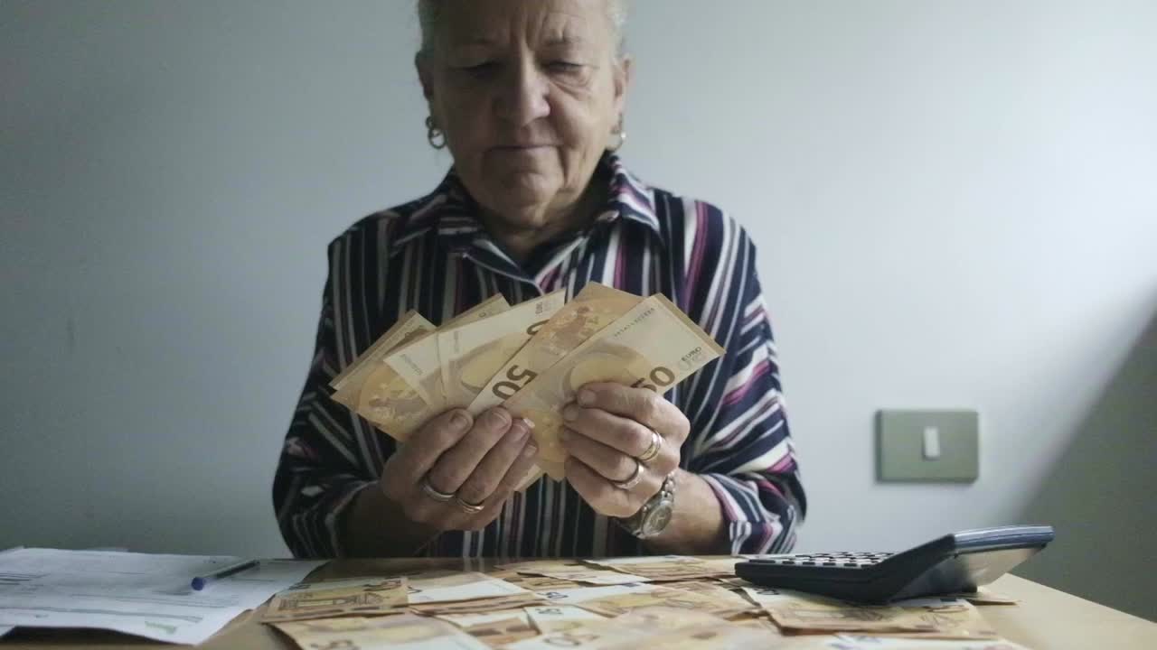 Donna caucasica di mezza età con capelli bianchi conta banconote da 50 euro, ufficio su scrivania con calcolatrice e tasse