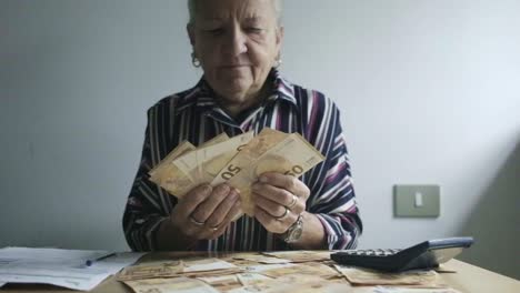 Middle-aged-caucasian-woman-with-white-hair-counts-50-euro-bills-paper-money,-desktop-office-with-a-calculator-and-taxes